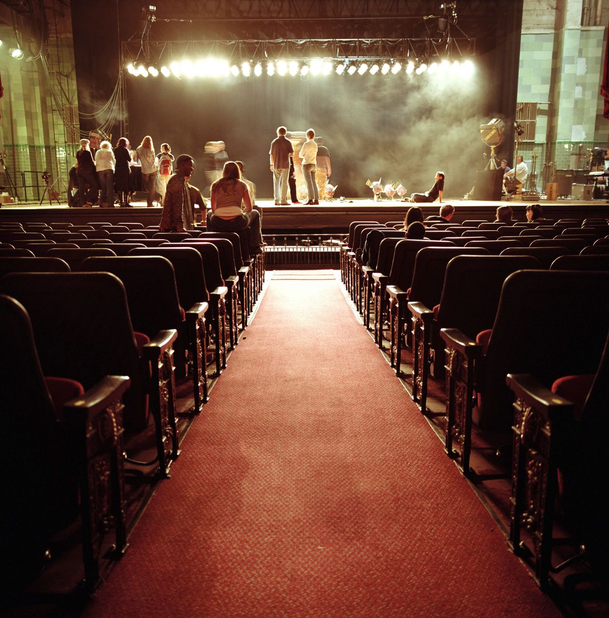 View down the aisle of an almost empty theatre auditorium towards the lit stage during a rehearsal